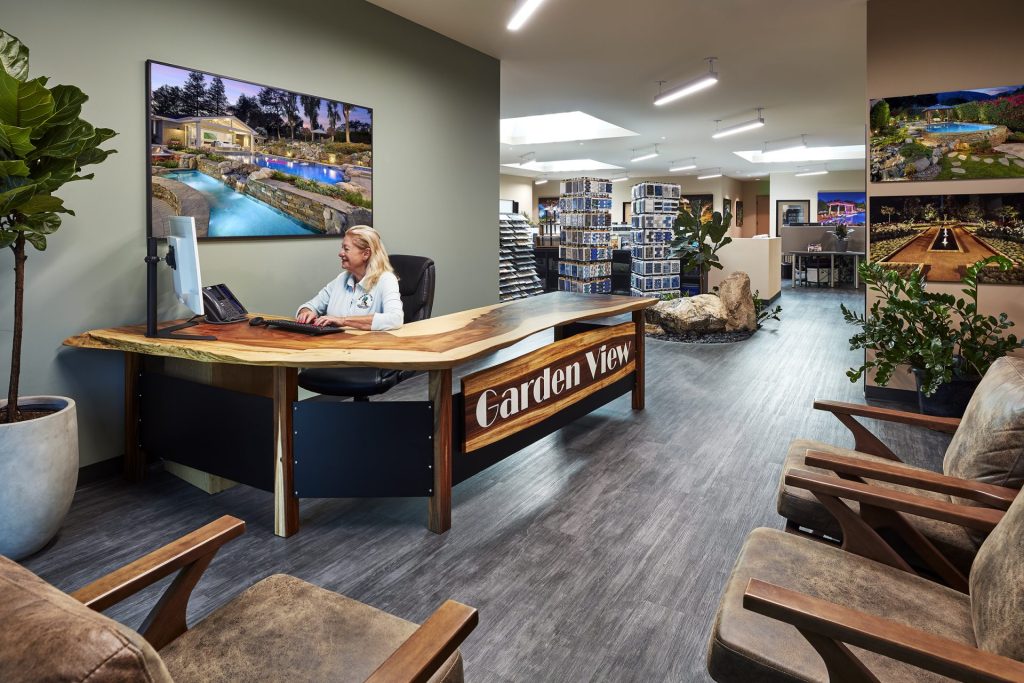 A woman sits at a wooden front desk labeled "Garden View" in a modern, well-lit office featuring lush plants and landscape-themed wall art. Comfortable chairs line the waiting area, with shelves and office spaces visible in the background.