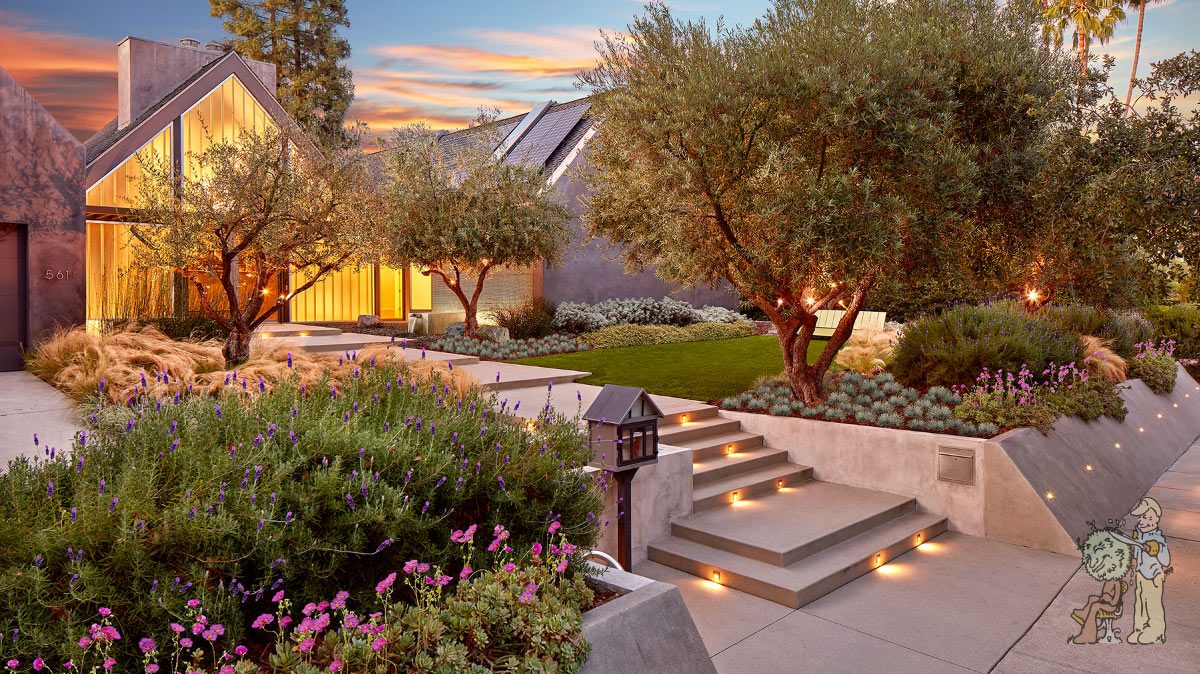 A modern house in Pasadena with angular rooflines is surrounded by a lush garden featuring a variety of plants and trees. Illuminated concrete steps lead up to the entrance, and evening lighting casts a warm glow across the landscape.