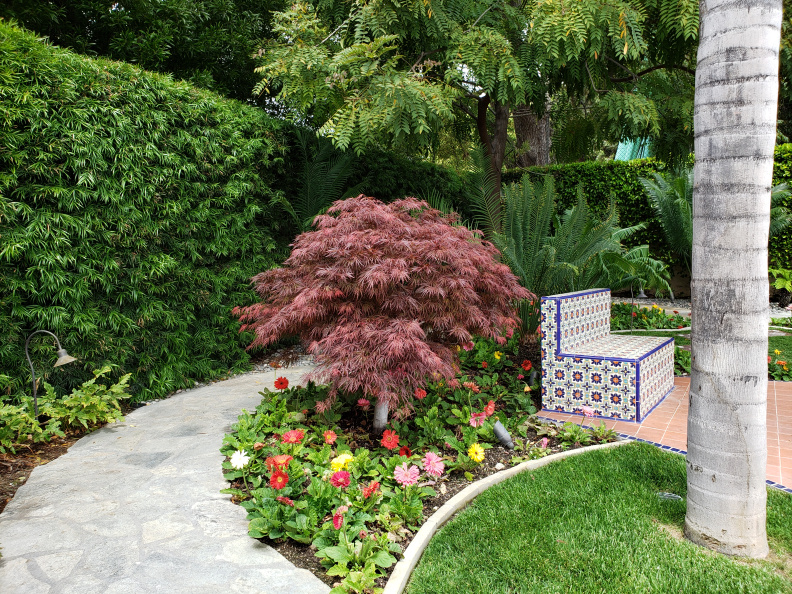 A curved stone path passes a small Japanese Maple tree and colorful flowers. Nearby is a mosaic tiled bench surrounded by lush green plants and palm trees.