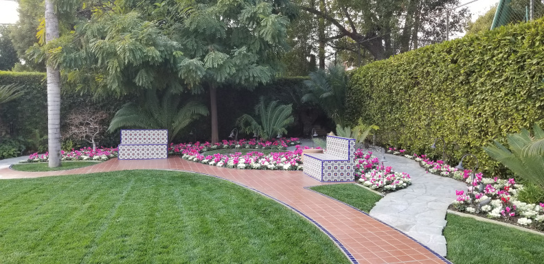 A So Cal garden with green grass, flowerbeds of pink and white flowers, tiled benches, a Mediterranean tile path, and tall hedges and trees in the background provide privacy.