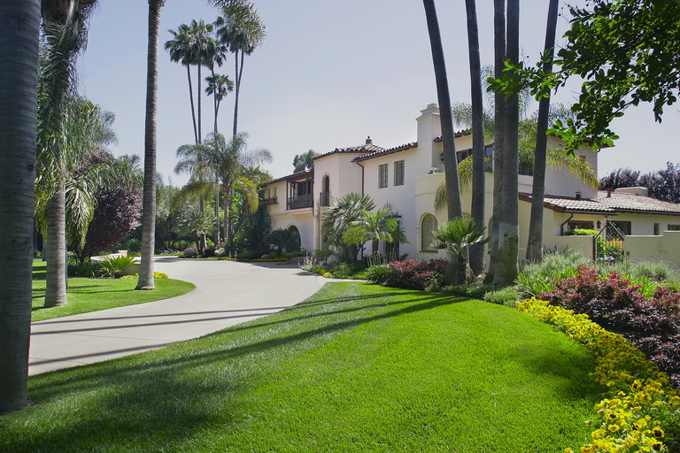 A large white Southern California house with Spanish-style architecture, surrounded by palm trees, a curved driveway, lush green lawn, and colorful flower beds under a sunny sky.